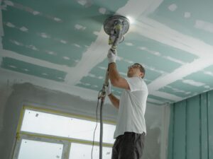 Painter using a dust-extraction sander to smooth a ceiling during popcorn ceiling removal